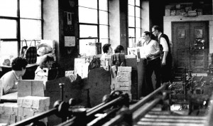 Women working at desks, 2 men chatting, machinery in forground